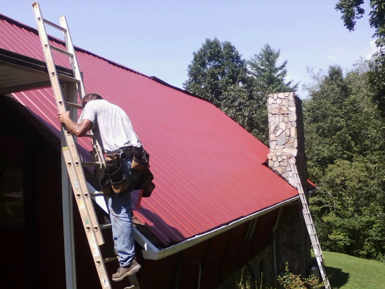 Roofer on a ladder beside a red metal roof and stone chimney — Soffit & Fascia Repair by Roofing Brothers in New Haven